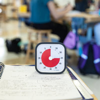 The Time Timer MOD 60 Minute visual timer is shown sitting on a notebook in a classroom setting. The red disk is set to 45 minutes and is shown as a three-quarters pie shape. 