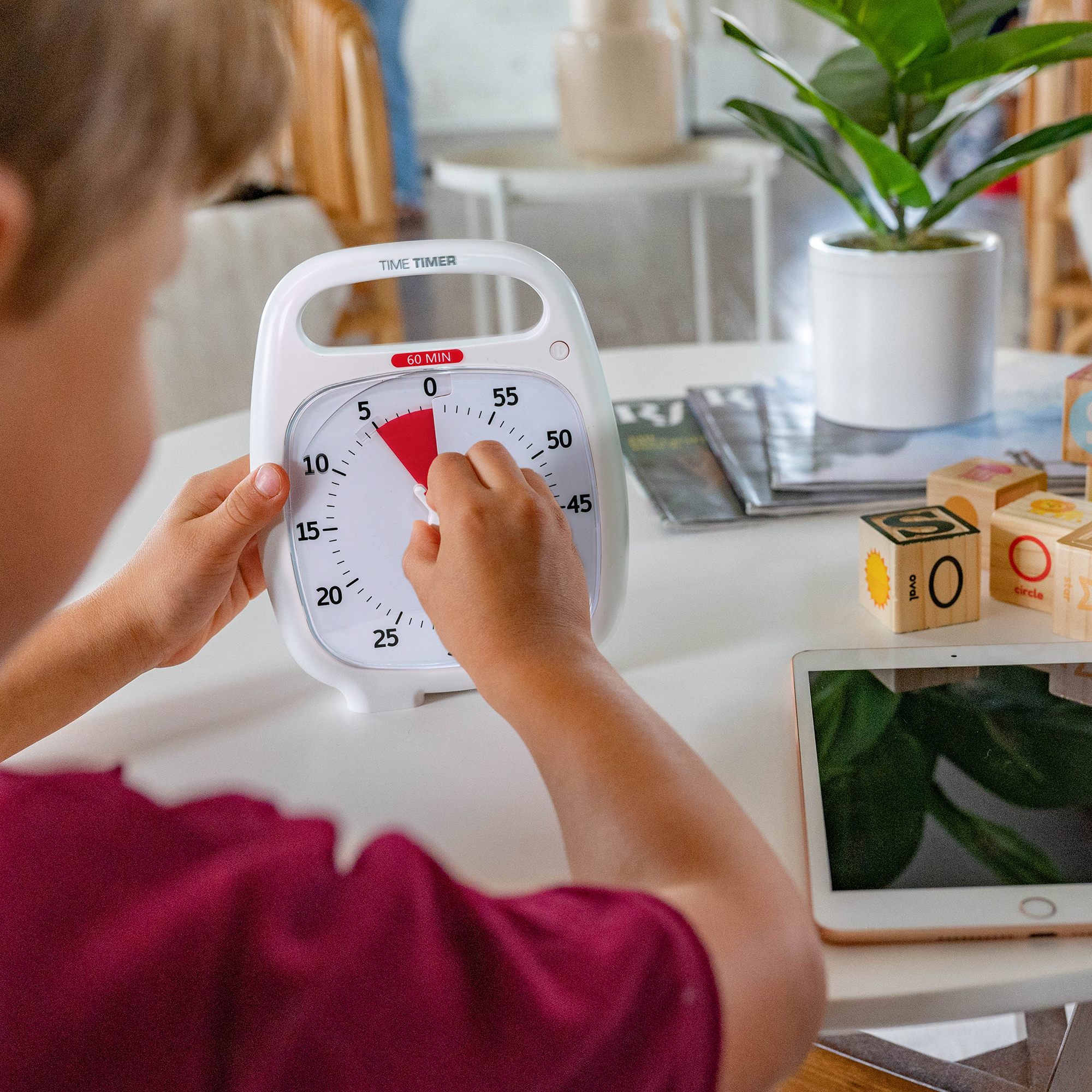A young boy is setting the Time Timer PLUS 60 Minute visual timer in white. He is turning the knob to set the red disk to 5 minutes. There is a tablet and blocks on the table. 