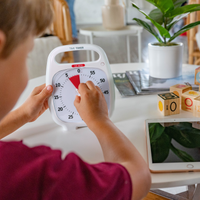 A young boy is setting the Time Timer PLUS 60 Minute visual timer in white. He is turning the knob to set the red disk to 5 minutes. There is a tablet and blocks on the table. 