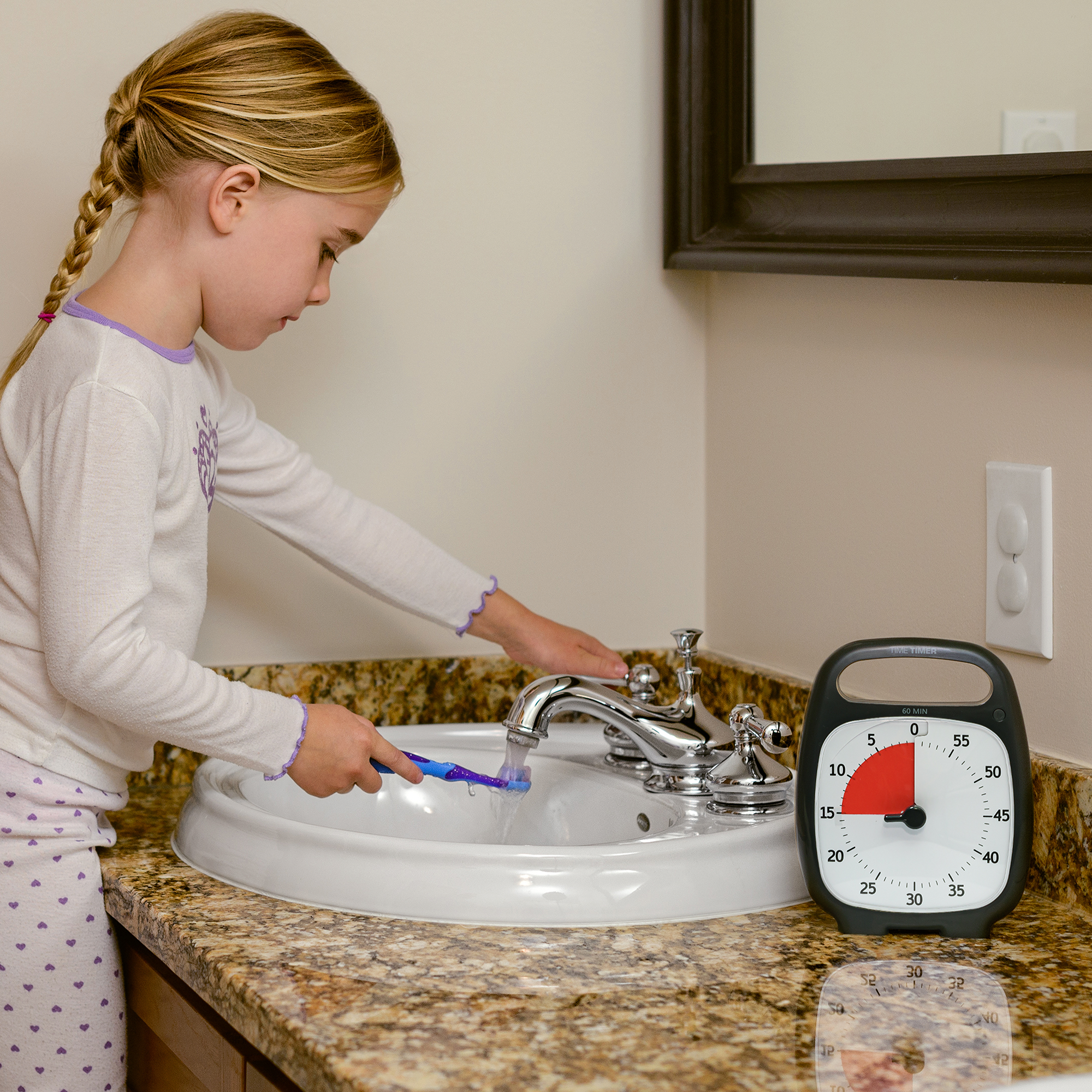 A young girl is standing at the bathroom sink wetting her toothbrush. The Time Timer PLUS 60 minute visual timer in Charcoal is sitting on the counter. The red disk of the timer is set to 15 minutes implying that she is using it for her bedtime routine. 