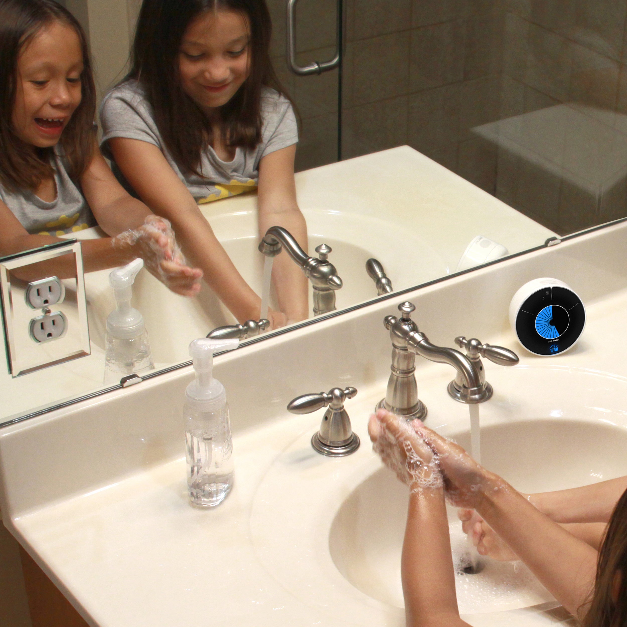Two young girls, ages 6 and 7, have fun and smile while washing their hands in a bathroom setting. The Time Timer WASH visual timer for handwashing sits on the counter beside the sink. 