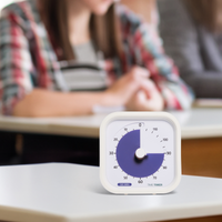 The Time Timer MOD - 120 Minute visual timer is shown sitting on a classroom desk. A student is shown in the background, out of focus. The calming purple disk of the timer is set to 90 minutes. 