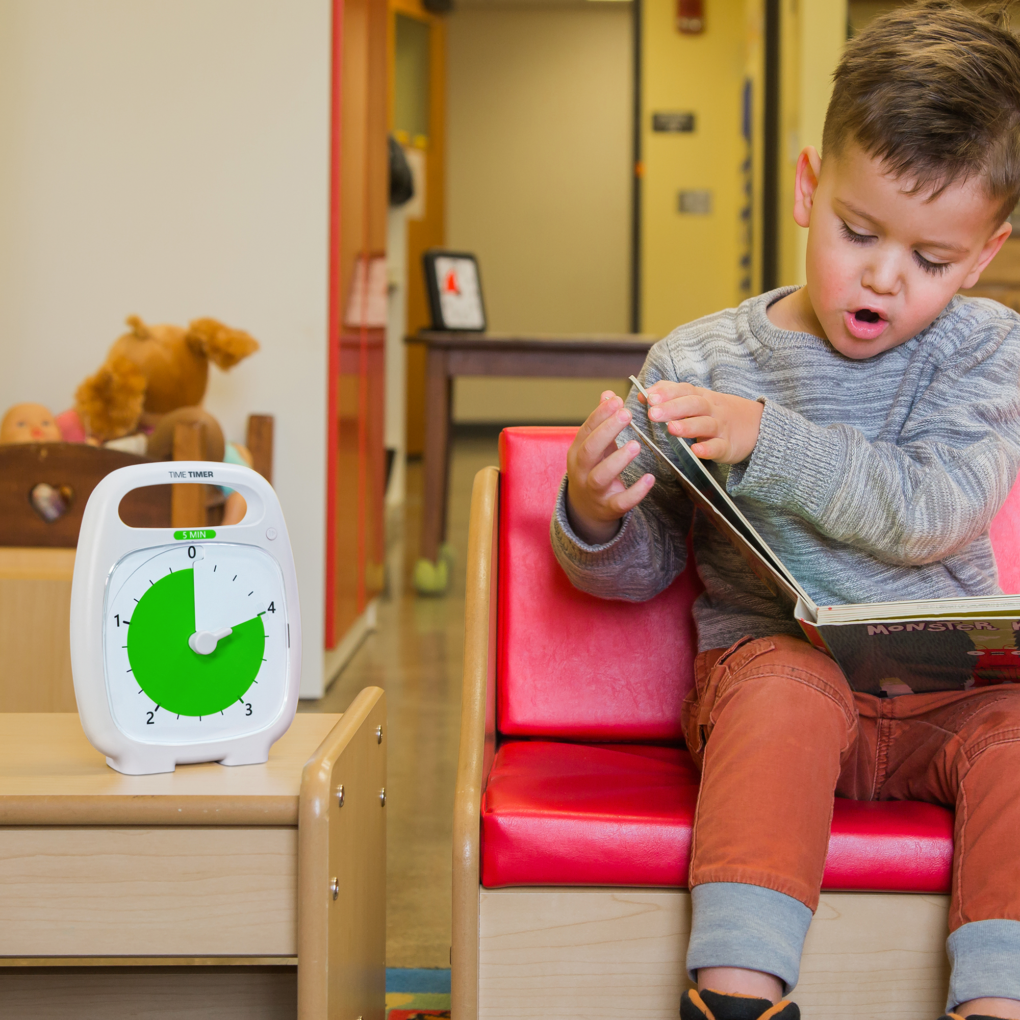 A young boy is sitting on a small couch in his preschool room, looking at a book. The Time Timer PLUS 5 minute visual timer is sitting on a small table next to him. The green disk of the timer is set to 4 minutes to show the child how much longer to spend on the book before moving to the next station.