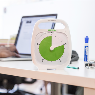 The Time Timer PLUS 5-minute visual timer is shown on a desk in an office setting. A laptop, pencil and dry erase marker are in the background. The green disk of the timer displays 4 minutes of time. 