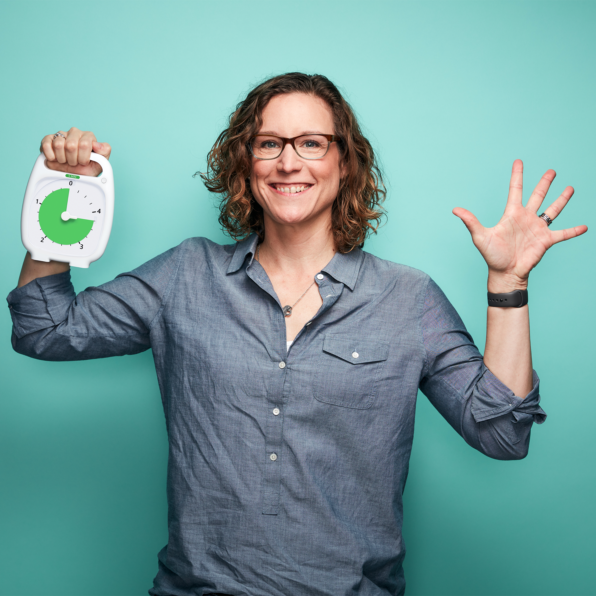 A smiling woman is standing in front of a light blue background. She is holding the Time Timer PLUS 5 minute visual timer in one hand, and holding up 5 fingers with her other hand. The green disk of the timer is displaying 3 minutes and 45 seconds.