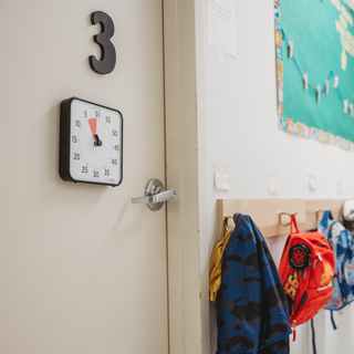 Children's classroom door with a Time Timer visual timer magnetically displayed showing 5 more minutes. This is displayed next to backpacks and coats.