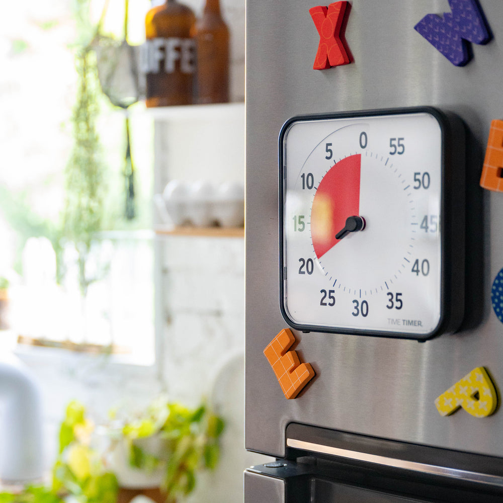 Time Timer attached to a refrigerator door with colorful magnetic letters around it. The timer is set to 15 minutes, with kitchen items visible in the background.