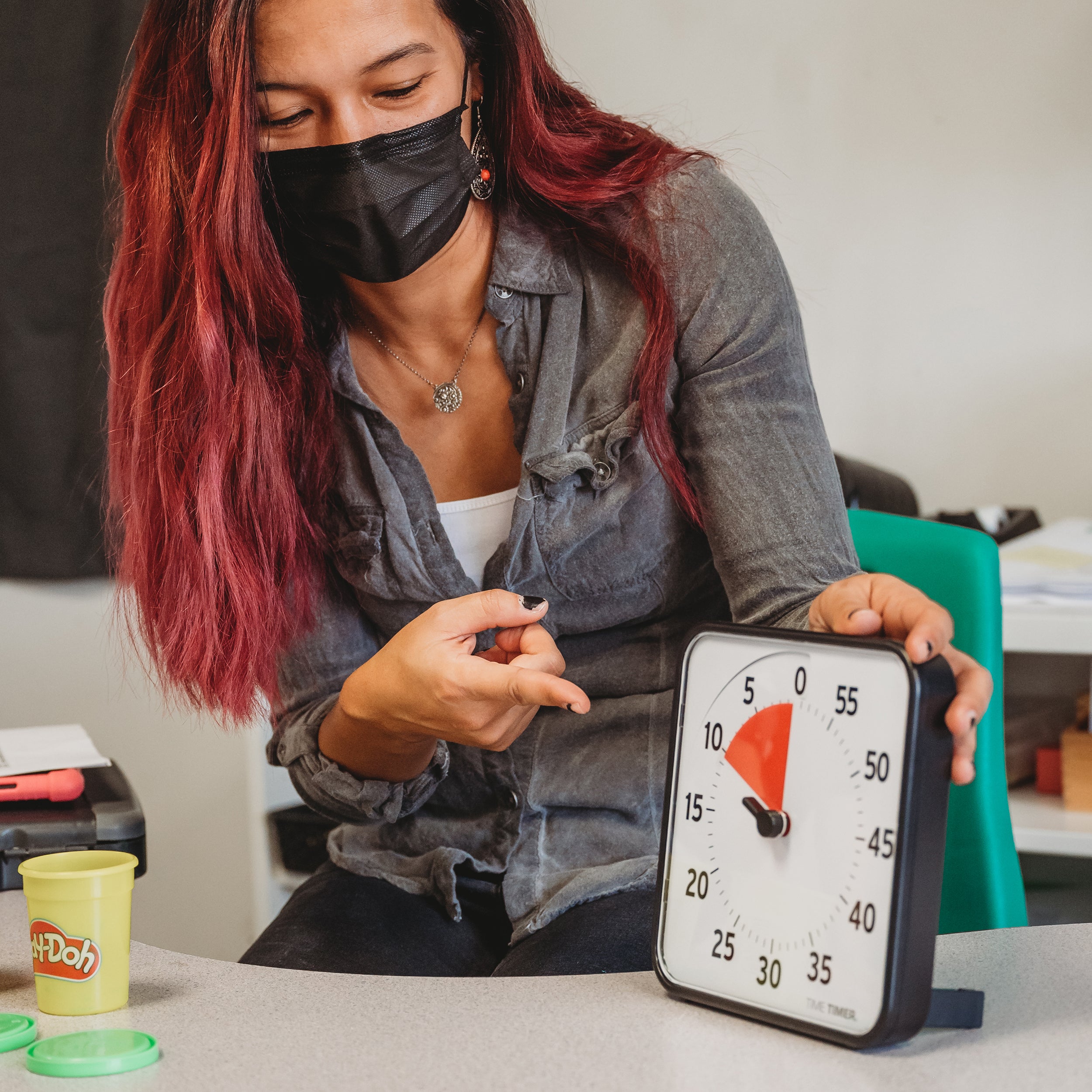 The Time Timer Original 8" visual timer is shown on the tabletop in a classroom. The teacher is pointing to the timer, which has the red disk set to 10 minutes. There is Play-Doh and other classroom supplies surrounding the teacher. 