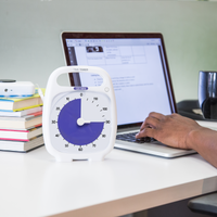 The Timer Timer PLUS 120 Minute visual timer is sitting on a desk in an office or home setting. Someone is using a laptop in the background and there is a pile of books sitting next to the computer. The purple disk of the Time Timer visual timer is set at 90 minutes to indicate a focused work or study session. 
