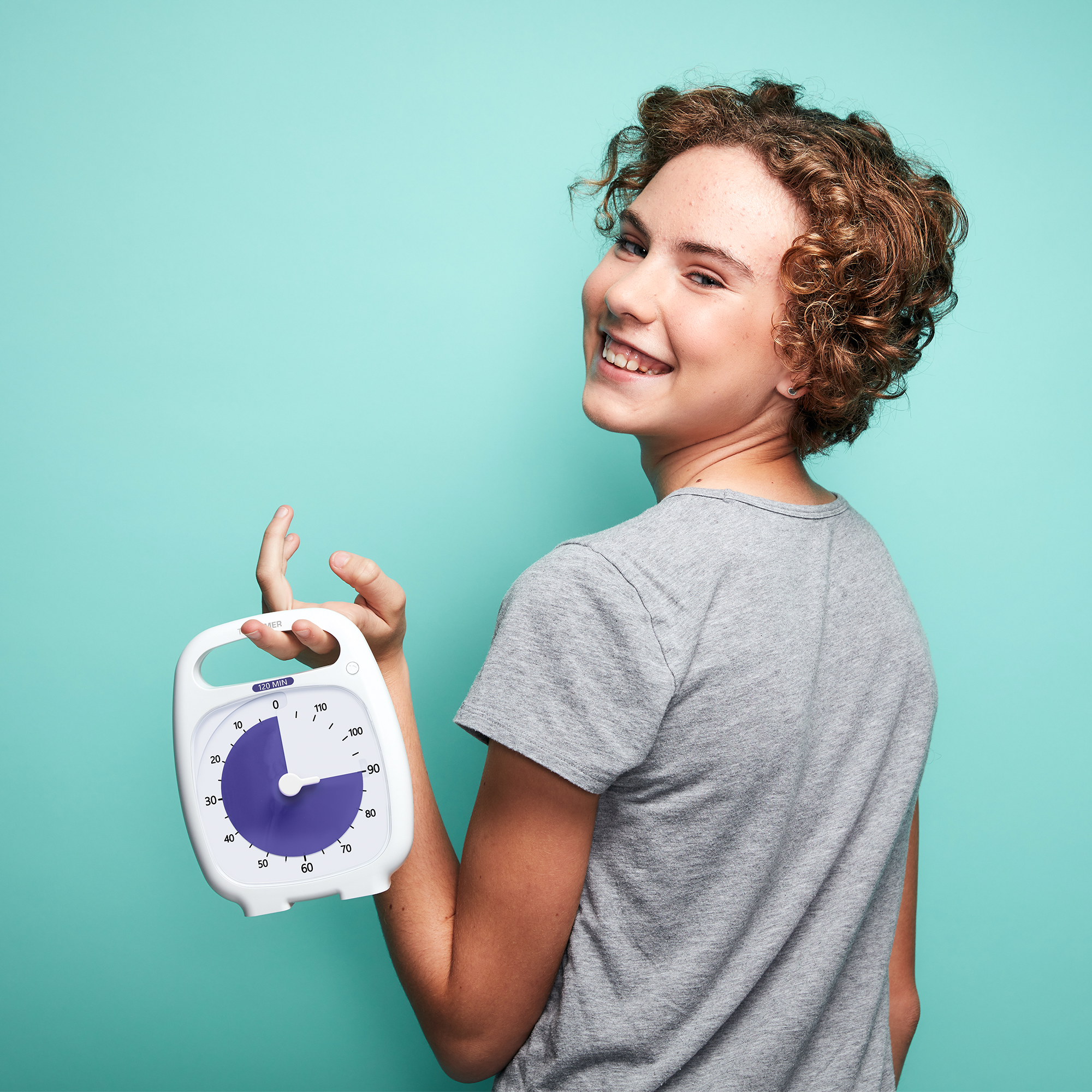 A smiling young woman with curly hair is standing in front of a bright blue background. She is holding the Time Timer 120 Minute timer. The purple disk is set to a 90 minute duration. 