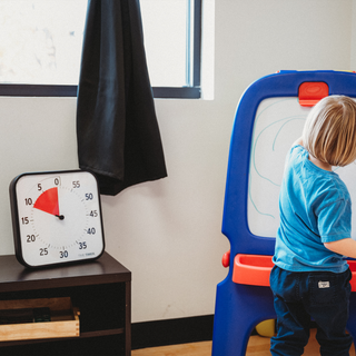 Child playing with a whiteboard in a classroom setting and a Time Timer visual timer sitting on a self next to him. It is showing about 10 more minutes of playtime. 