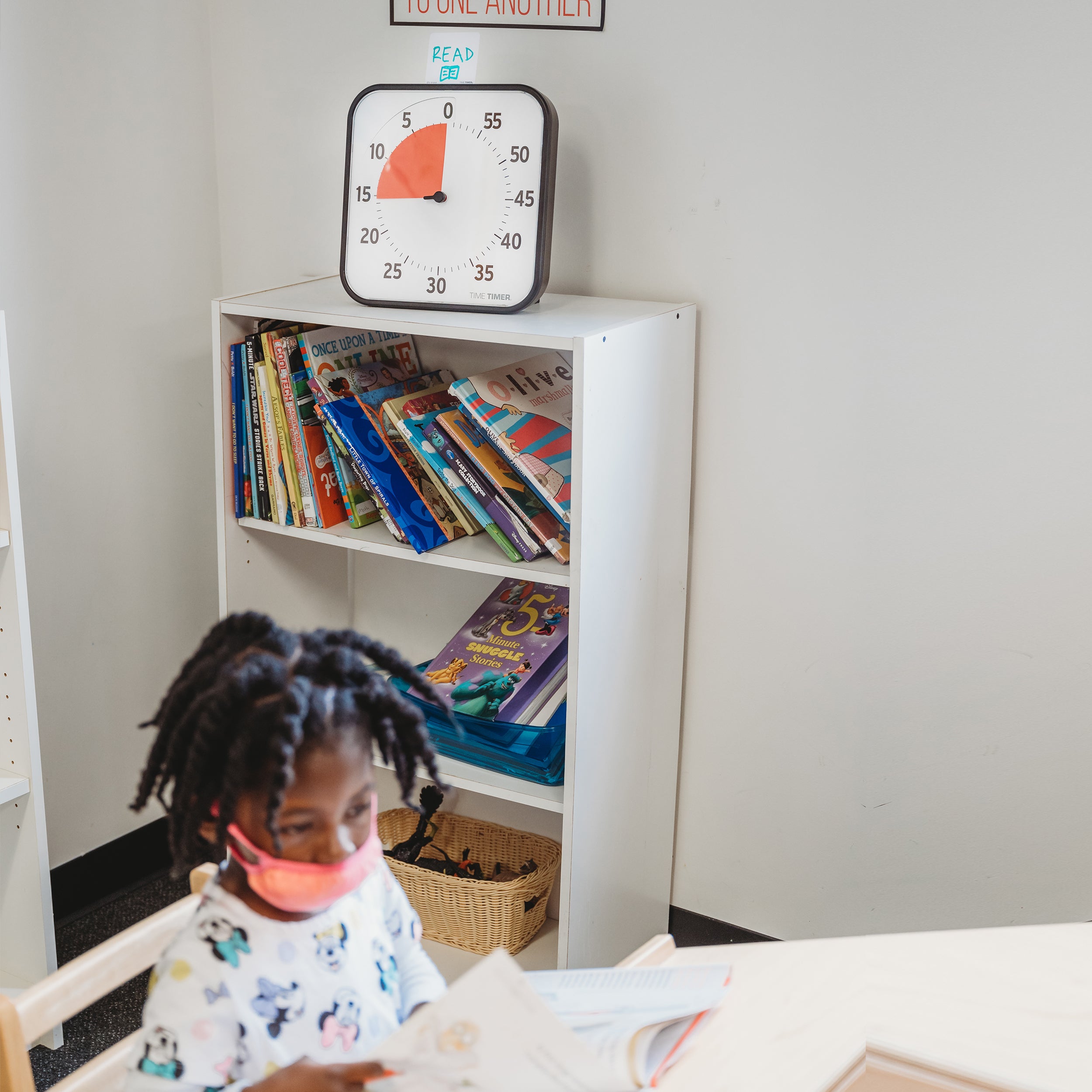 The Time Timer Original 12" visual timer sits on the top of a 3-tier bookshelf. The included Dry Erase Activity Card is placed on top of the timer with "Read" written on it along with a drawing of a book. A child sits at the desk in front of the bookshelf reading a book.