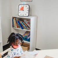 The Time Timer Original 12" visual timer sits on the top of a 3-tier bookshelf. The included Dry Erase Activity Card is placed on top of the timer with "Read" written on it along with a drawing of a book. A child sits at the desk in front of the bookshelf reading a book. 