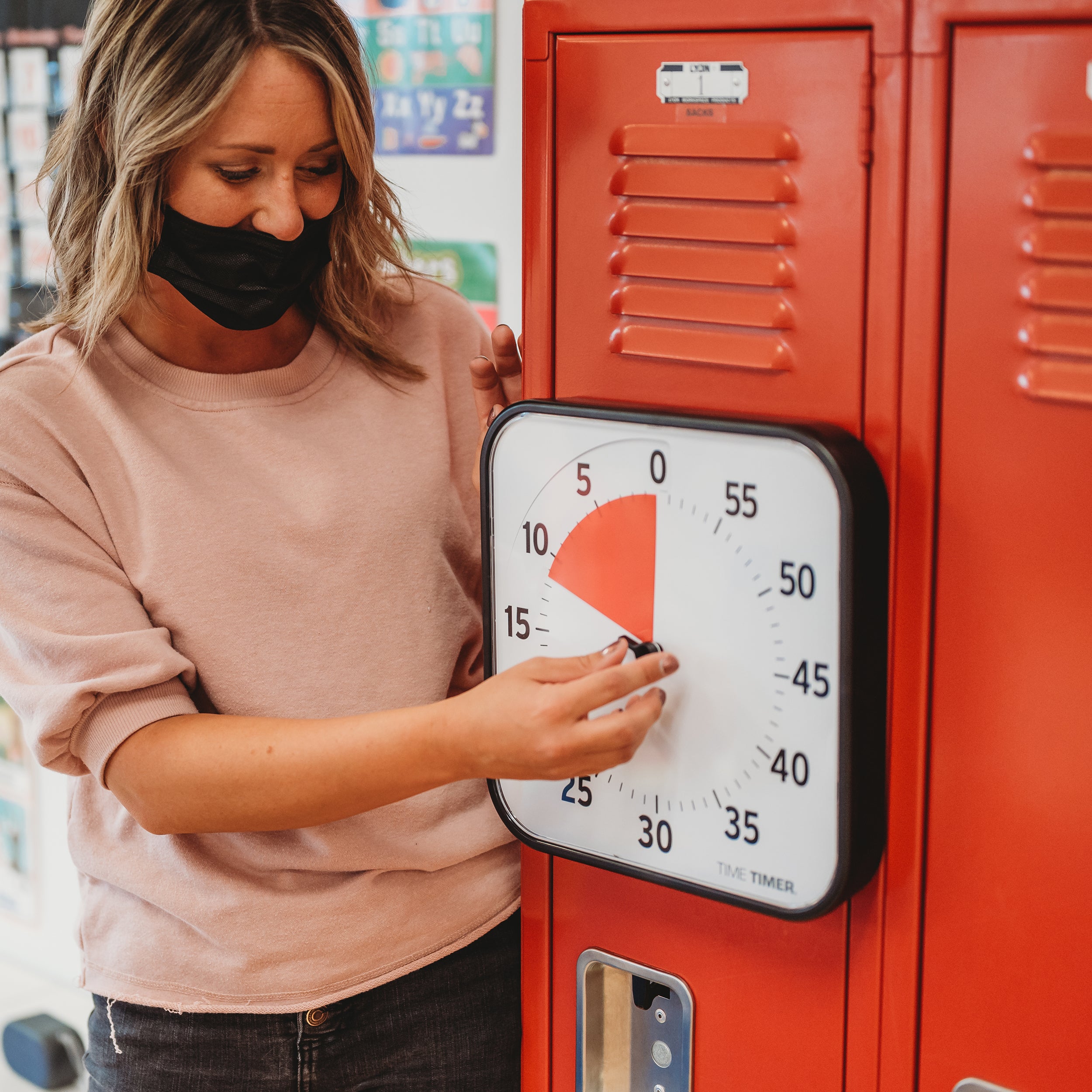 The Time Timer Original 12" visual timer is shown magnetically sticking to red lockers. A teacher is using the center dial of the timer to set the duration, around 10 minutes.