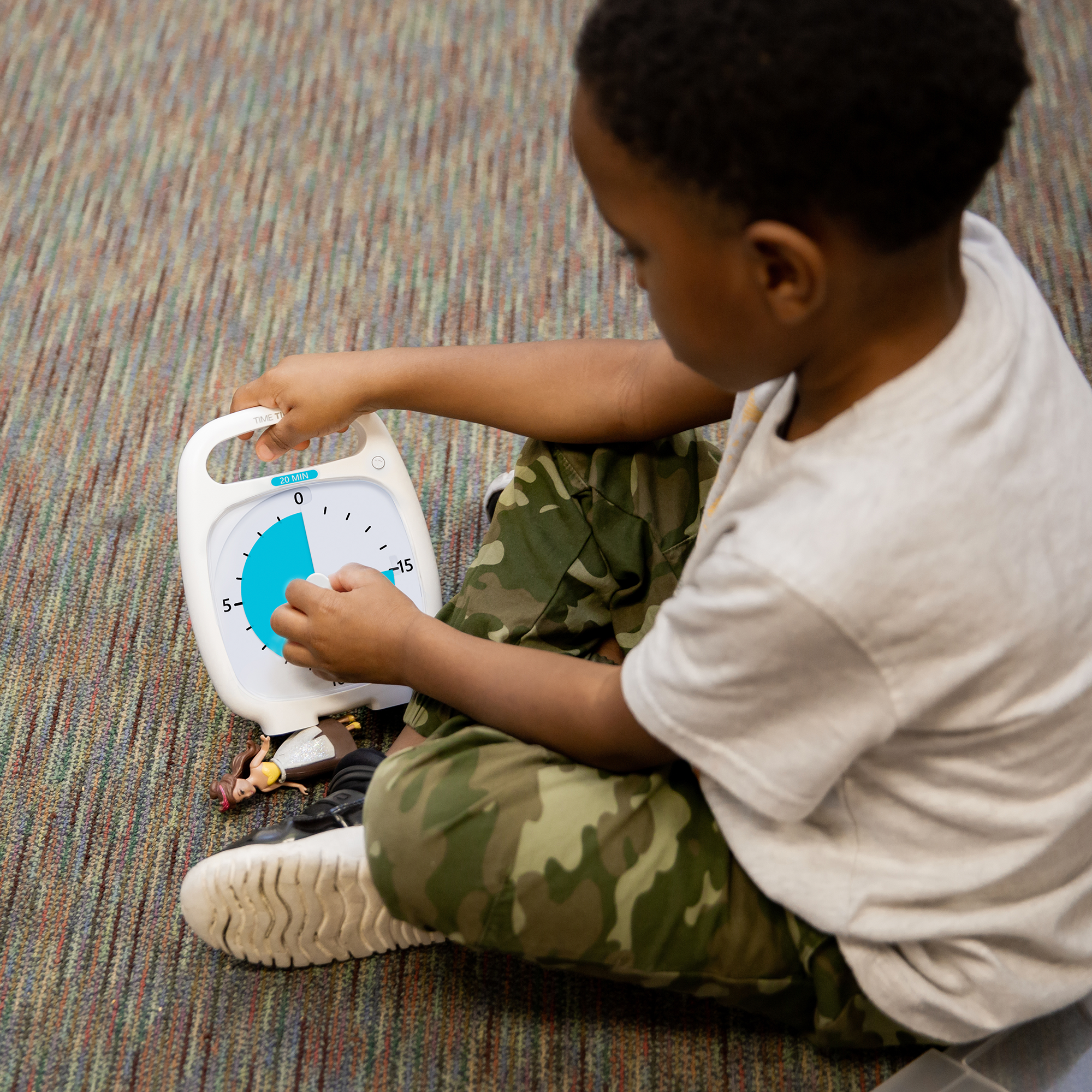 A young boy sits on the floor next to a small doll. He is setting the Time Timer PLUS 20 Minute Visual Timer by using the center knob. He is turning the duration to about 15 minutes. 