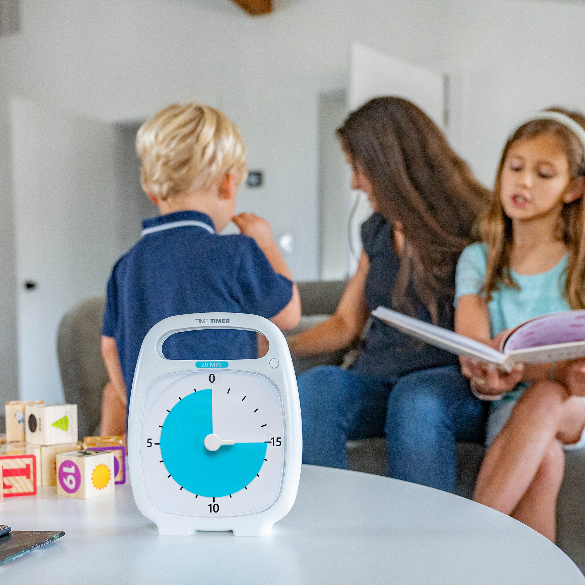 A Time Timer PLUS 20 minute visual timer is sitting on a table in a living room. It's blue disk is set to 15 minutes. In the background, a mom and two kids are on the couch, reading books and talking. There a play blocks sitting on the table next to the timer.