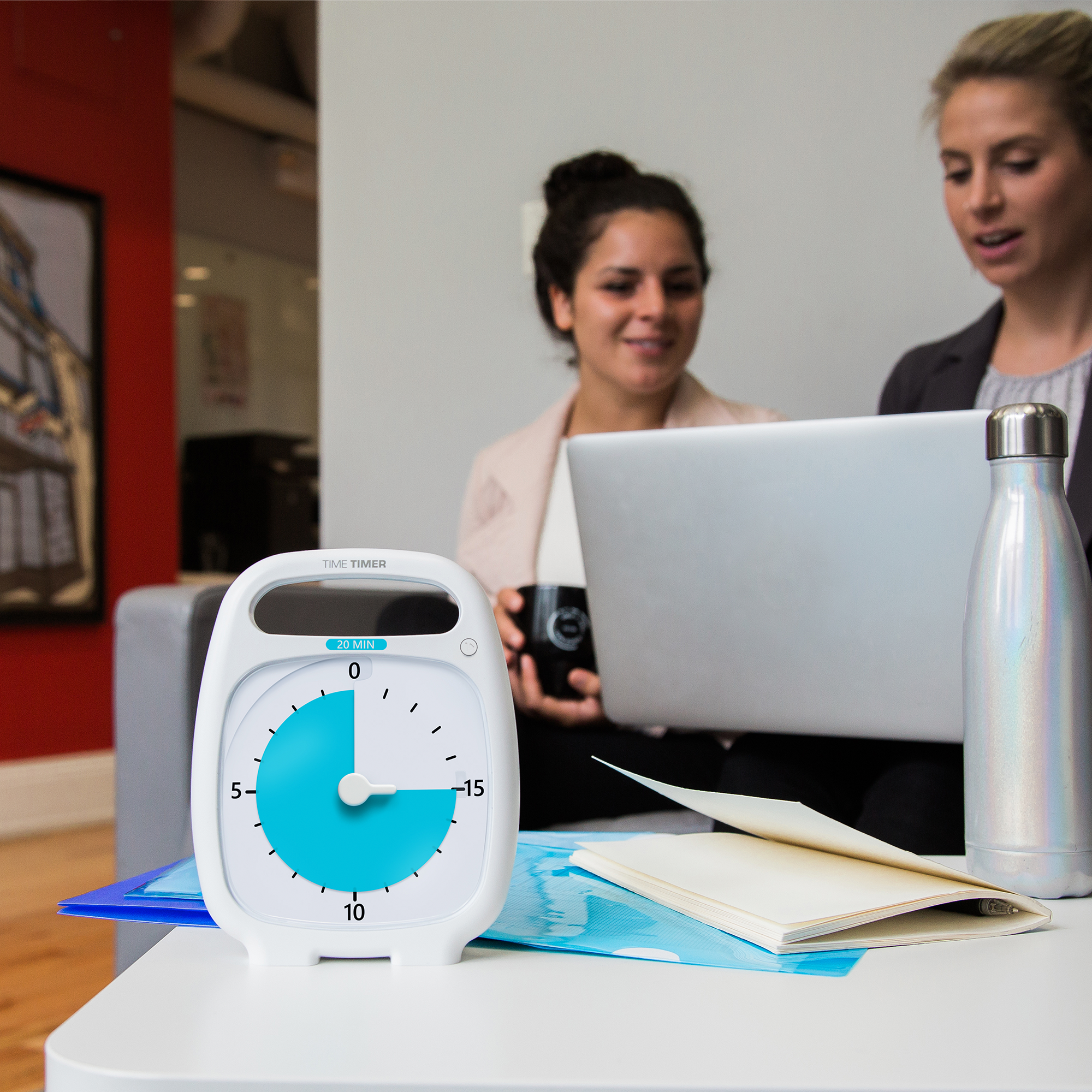 The Time Timer PLUS 20 Minute visual timer is sitting on a table in an office setting. The bright blue disk is set at a 15 minute duration. Next to the timer, on the table is a water bottle and a notepad. In the background are two women sitting on a couch, talking and looking at a laptop. 