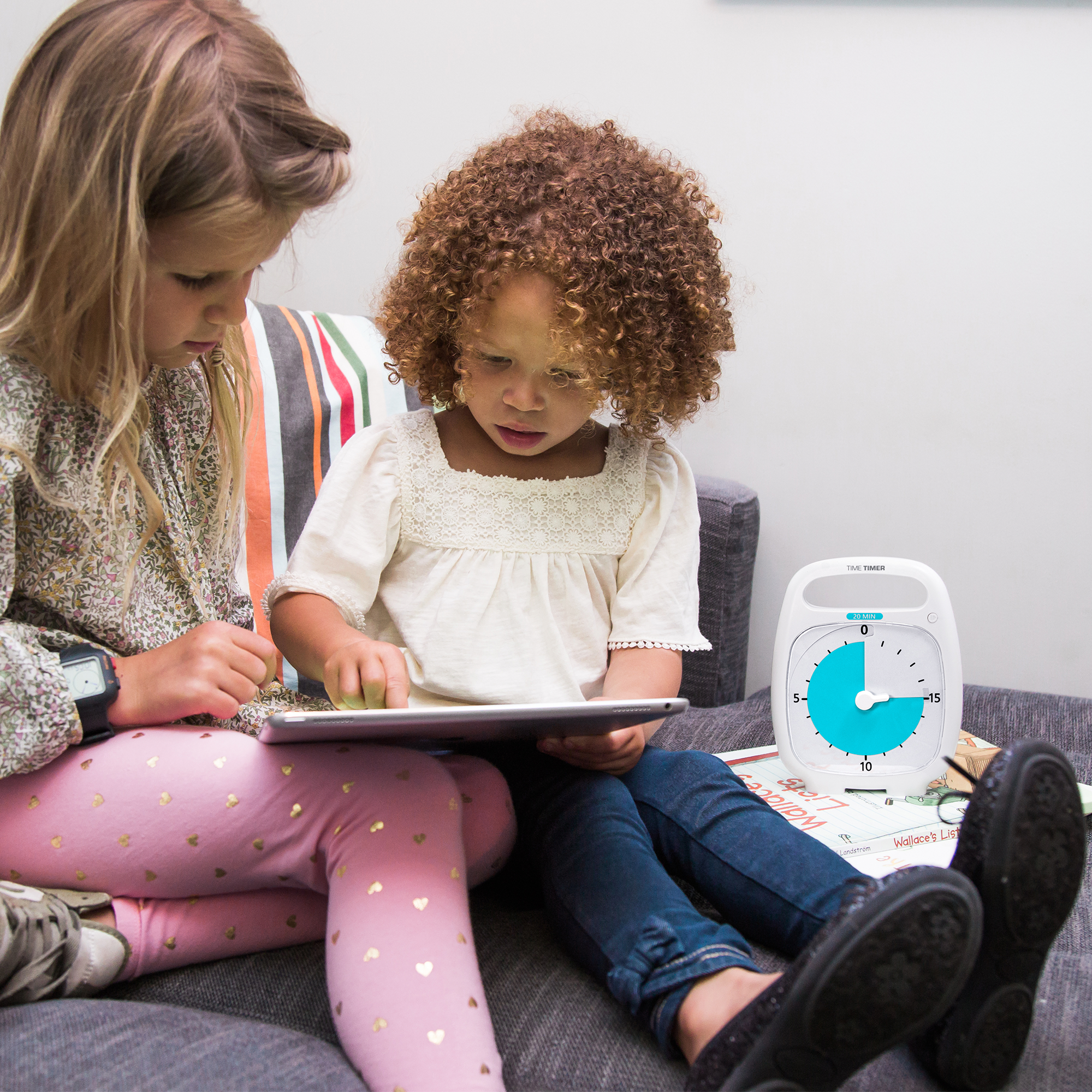 Two young girls are sitting on a couch reading together. The Time Timer PLUS 20 Minute visual timer is placed on the couch next to them. The bright blue disk is set at a 15 minute duration. 