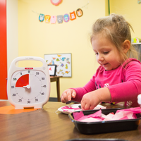 A young girl is making a craft at a table in her preschool. The Time Timer PLUS 60 minute visual timer is placed on the table next to her. The red disk is st to 15 minutes.