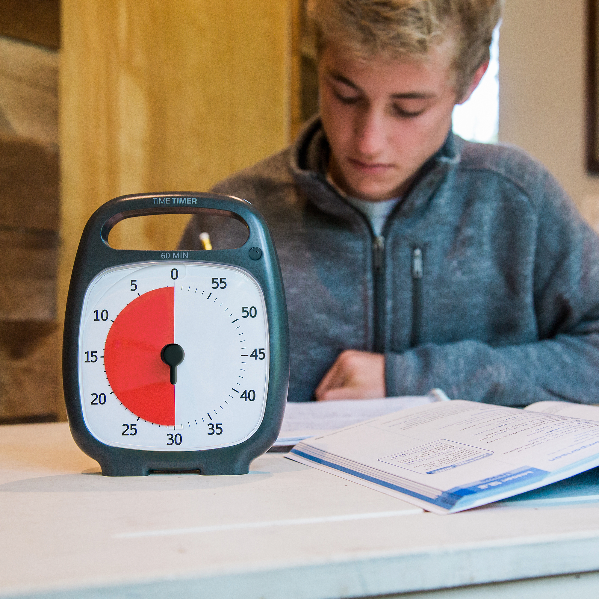 A young man is sitting at a table doing homework. The Time Timer PLUS 60 minute visual timer in the charcoal color is sitting in front of him. The red disk on the timer is set to 30 minutes. 