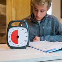 A young man is sitting at a table doing homework. The Time Timer PLUS 60 minute visual timer in the charcoal color is sitting in front of him. The red disk on the timer is set to 30 minutes. 