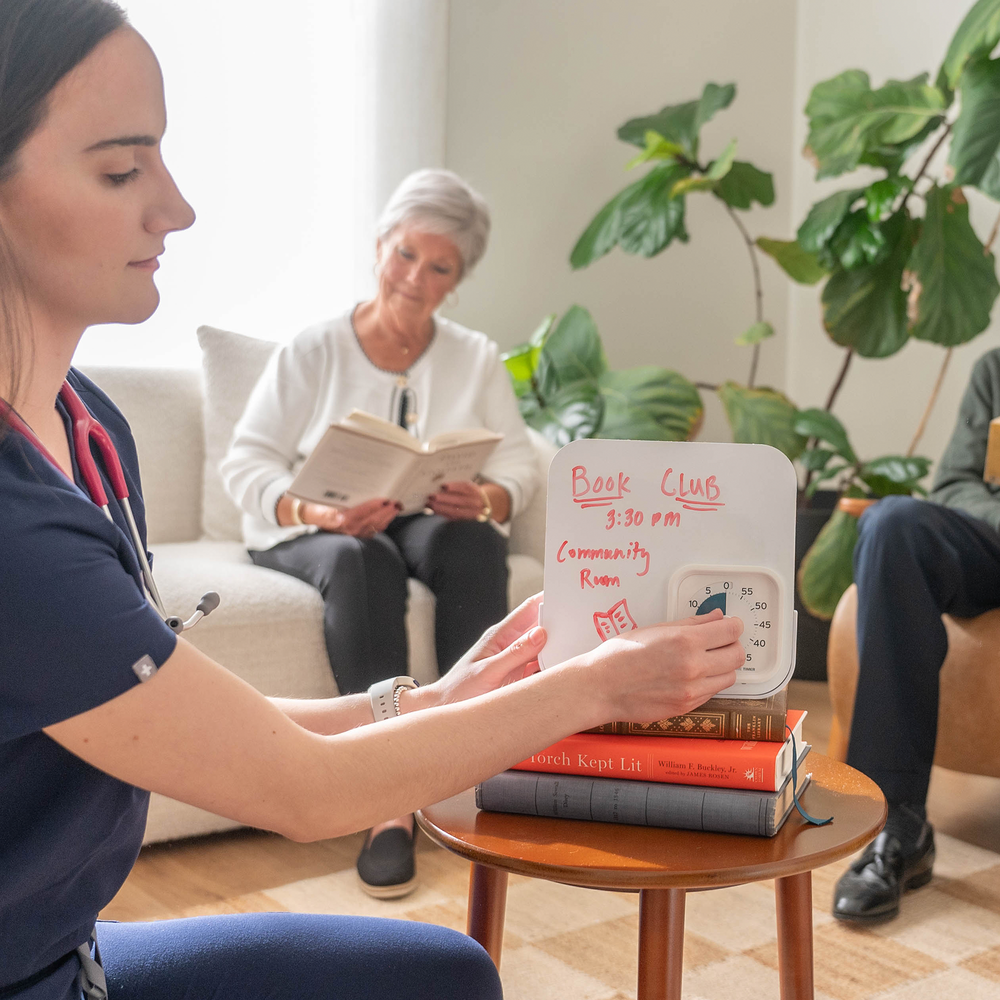 Woman holding a book club sign with others in the background in a cozy living room.