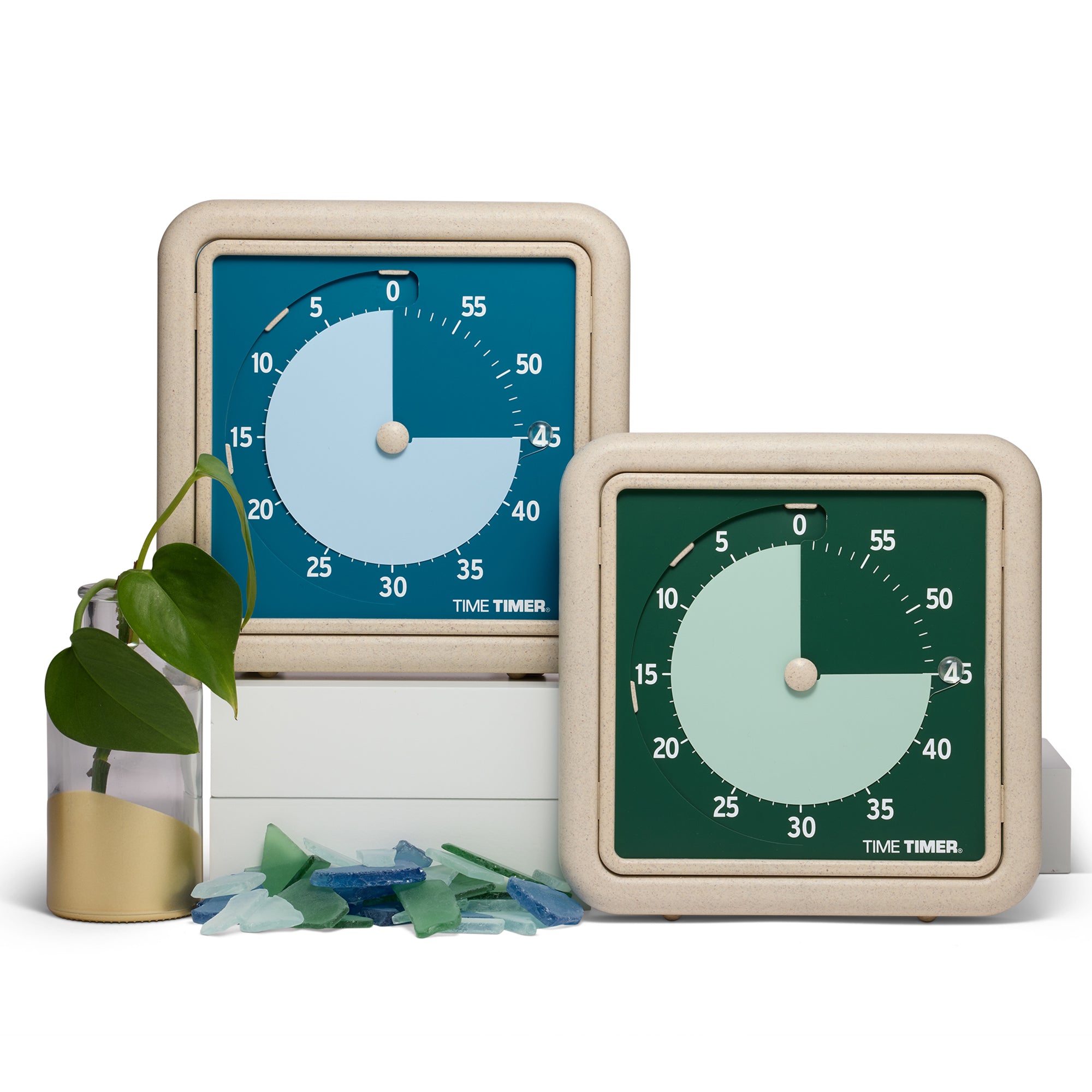 Two 60-minute visual timers are sitting next to each other. One is a dark blue color with light blue disk. The other is a dark green color with light green disk. Both have a textured beige frame. They are placed next to a green plant in a glass and gold vase and pieces of sea glass are scattered in front. 