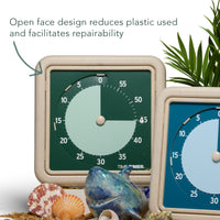 The Time Timer RETRO - Eco Edition 60-minute visual timers are shown along with some plants, sea shells, rice husks, and a shark figurine. An arrow is pointing to the timer with a message stating "Open face design reduces plastic used and facilitates repairability." This indicates that there is no lens covering the face of the timer, therefore using less plastic. 