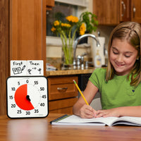 The Time Timer® Original 8” visual timer is shown on a table next to a young child doing homework. The red disk of the timer is set to 35 minutes. There are two Dry Erase Activity Cards placed on top of the timer. The first one shows a drawing of a book and pencil with the word "First" written. The second card shows a picture of 2 stick figures tossing a ball with the word "Then." Together, these two cards show an example of a First/Then schedule.