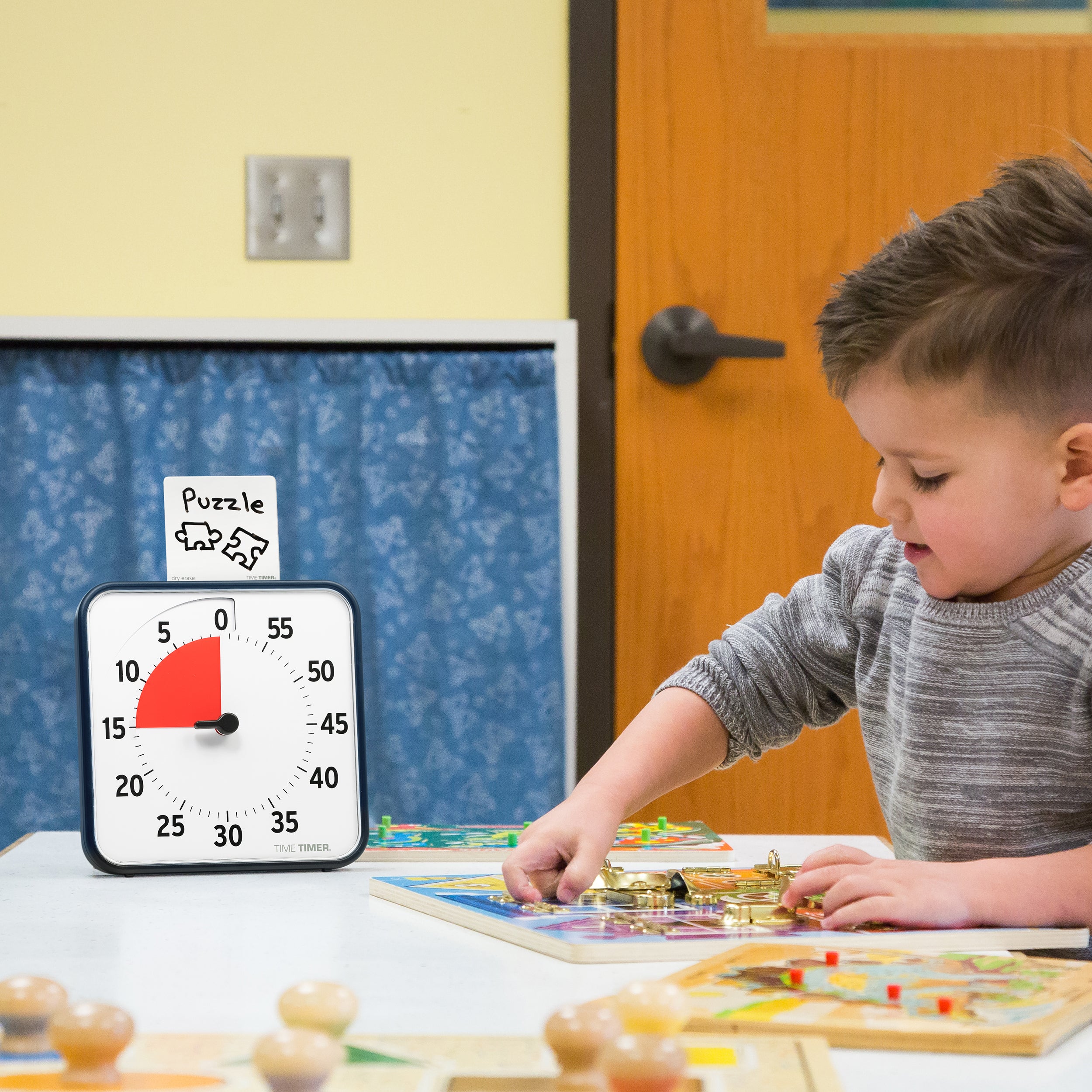The Time Timer® Original 8” is shown on a tabletop in the classroom. A young child of preschool age is playing with puzzles. The visual timer has a red disk set to 15 minutes. In the top of the timer, the Dry Erase Card accessory is shown with the word "Puzzle" written on it and a drawing of two puzzle pieces. 