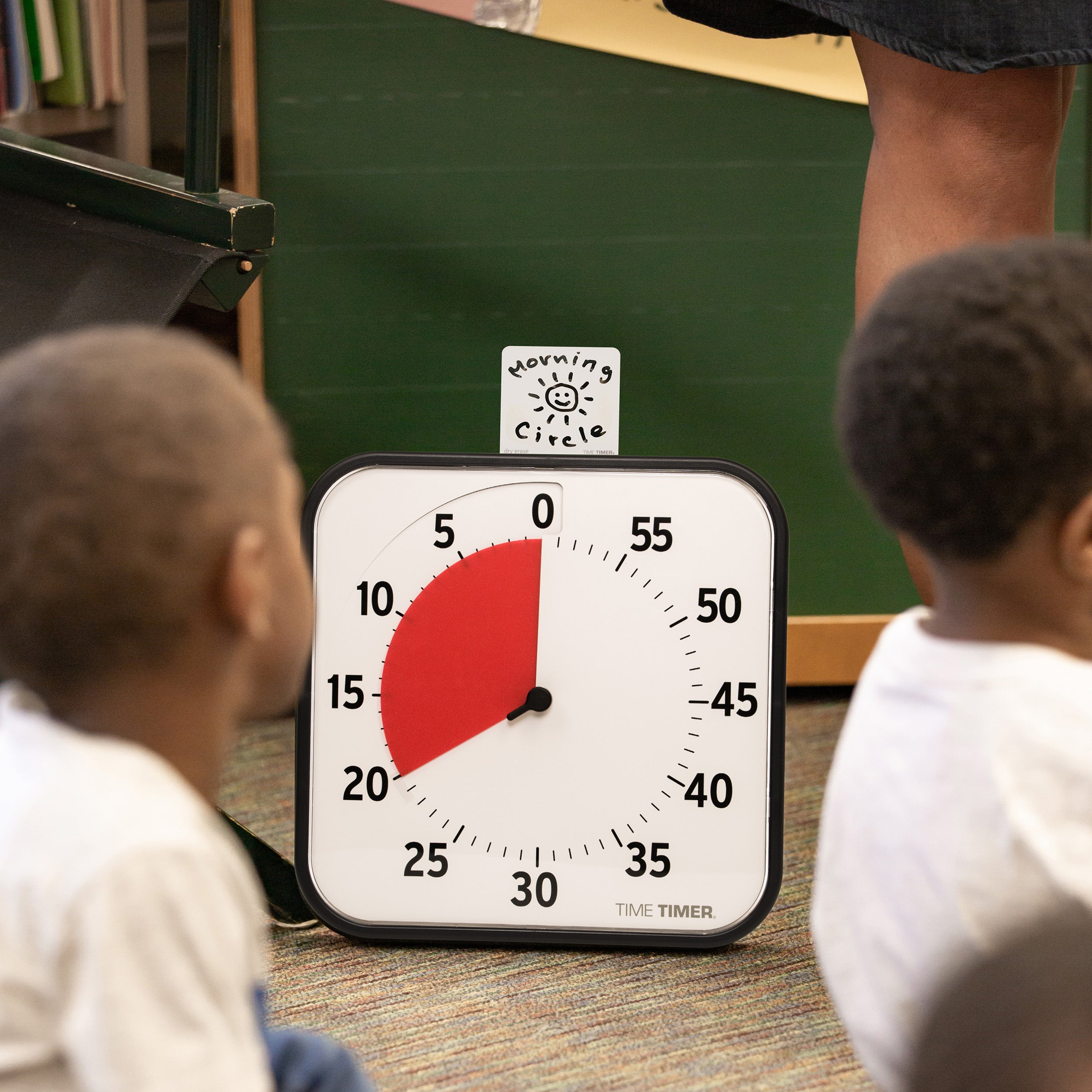 The Time Timer Original 12" is shown on the floor of a classroom. Drawn on the Dry Erase Card Accessory is "Morning Circle" with a sunshine. Children are sitting around the 60-minute visual timer.