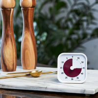 Time Timer MOD - Home Edition - This sophisticated visual timer is pictured in a kitchen setting with a marble serving tray, brass measuring spoons and wooden salt and pepper shakers. It is a white timer with a rich burgundy colored disk that disappears as time elapses. 