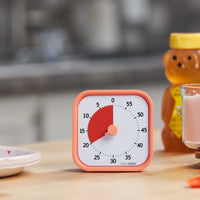 Time Timer MOD - Home Edition - This energetic visual timer is pictured on a kitchen table with a bear-shaped container of honey and some fun paper plates in the background. This is the Dreamsicle Orange color and has a sherbert colored exterior with a saturated orange colored disk that disappears as time elapses.