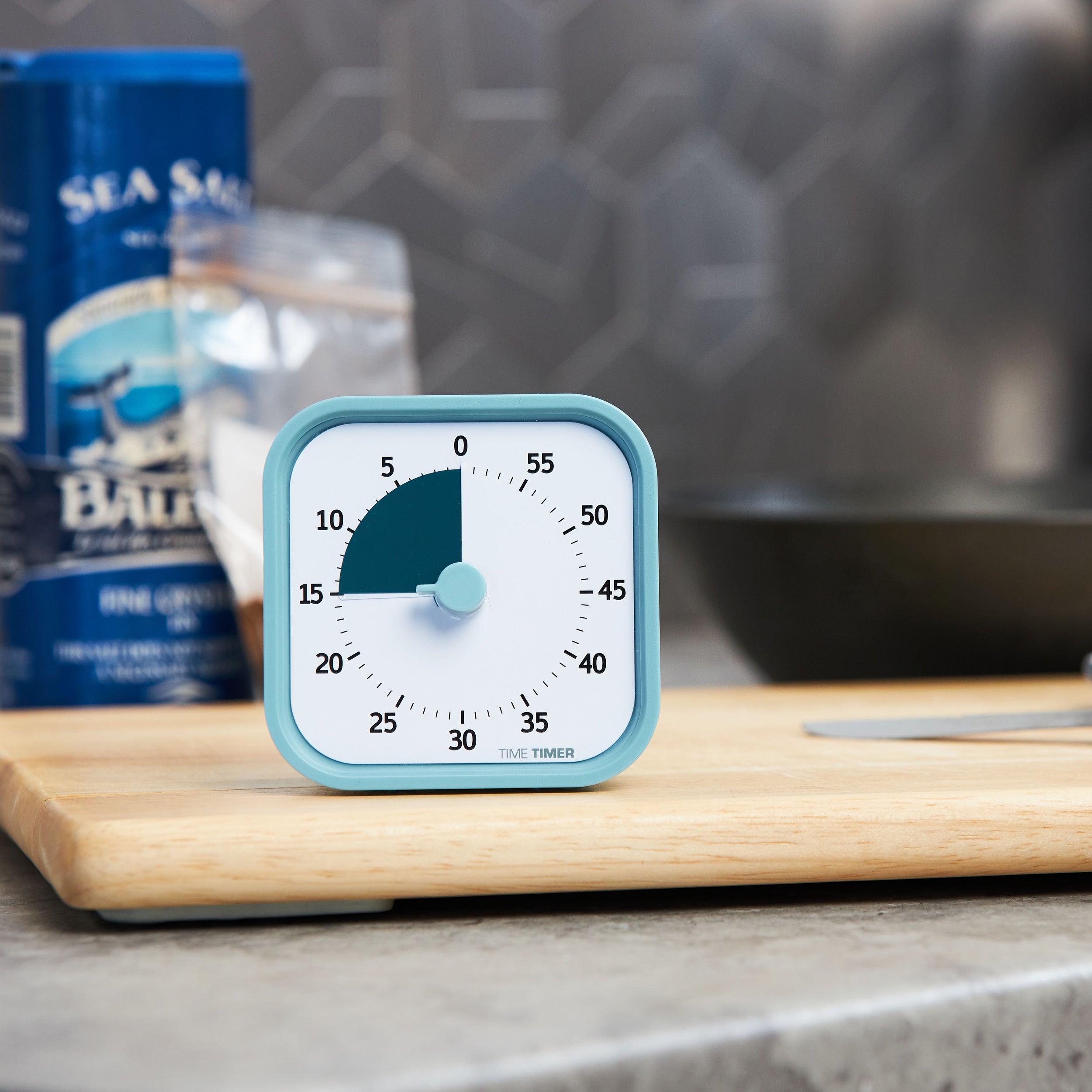 Time Timer MOD - Home Edition - This relaxing visual timer is pictured on a kitchen countertop atop a wooden cutting board with a container of salt, a baggie of spices and a bowl in the background. This is the Lake Day Blue color and has a pale blue exterior with a dark teal-blue colored disk that disappears as time elapses.