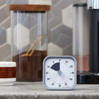 Time Timer MOD - Home Edition - This calming visual timer is pictured on a kitchen countertop with a coffee mug and canister of coffee ground in the background. This is the Pale Shale color and has a light grey exterior with a dark grey-purple colored disk that disappears as time elapses.